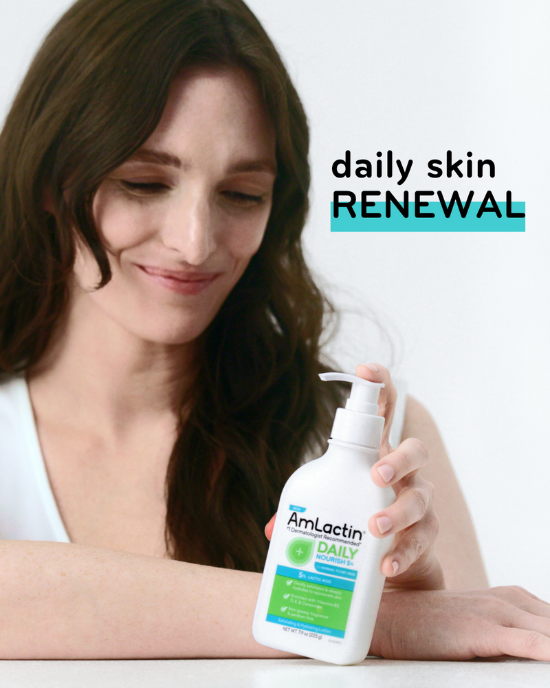 Smiling dark-haired woman in a white tank top on a light grey background. She is resting her one arm on a white table and holding AmLactin Daily Nourish 5% Pump Top Lotion bottle in her other hand.