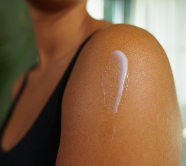 Close-up of a person's shoulder with a streak of white skincare product or sunscreen applied to their skin, wearing a black tank top against a softly blurred background.