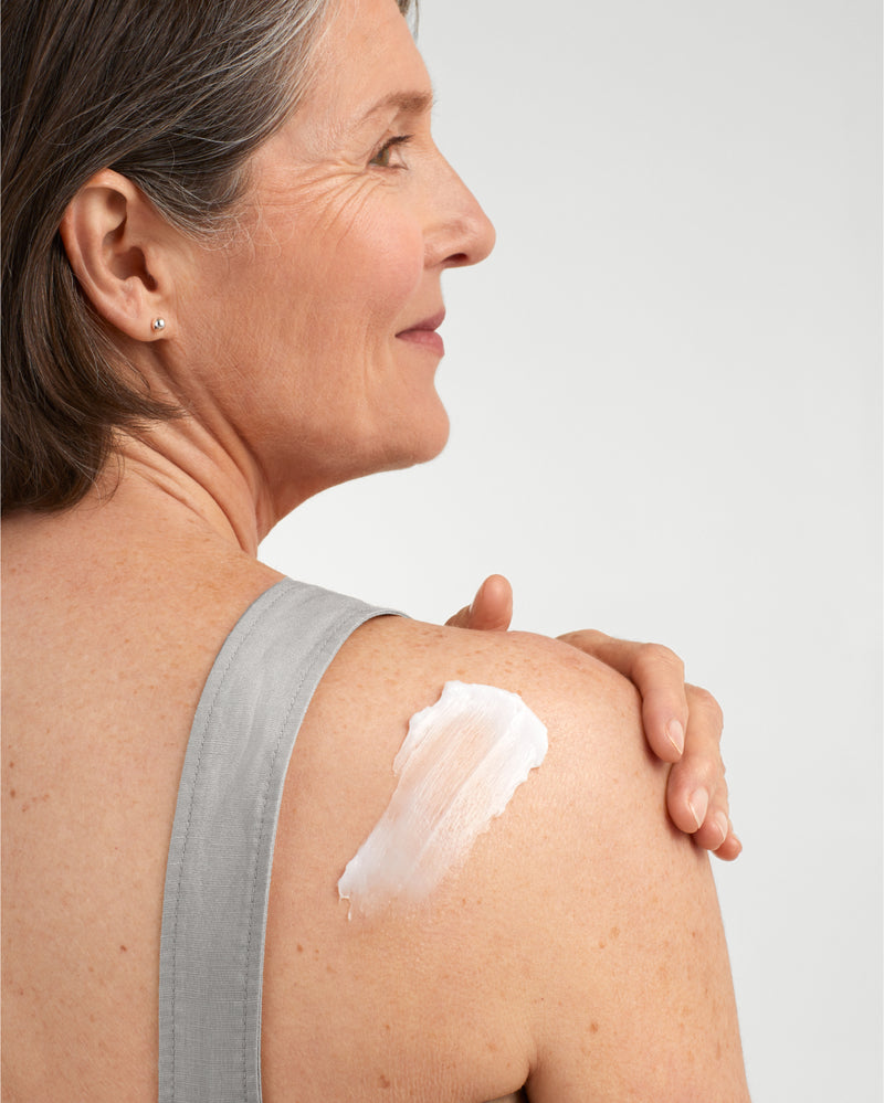 Mature, grey-haired woman in a medium grey tank top with her back and shoulder visible. Shoulder has a swatch of AmLactin Crepe Firming cream on it.