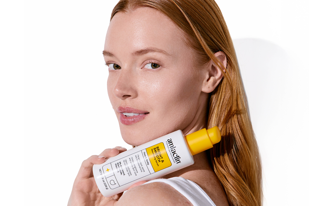 A woman with long red hair holds a white bottle of Amlactin against her neck on a clean white background.
