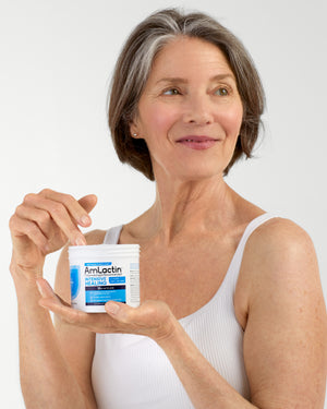 Mature grey-haired woman in white tank top on a light grey background holding AmLactin Intensive Healing Cream Tub in the palm of one hand. Her other hand is dipping into the tube for cream.