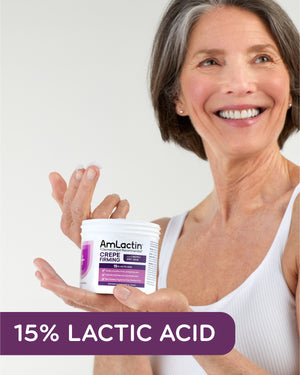 Older White Female in White Tank Top Holding AmLactin Crepe Firming Tub in Her Hand, Cream on Two Fingers of Other Hand. 15% Lactic Acid Flag in purple across bottom of the image.