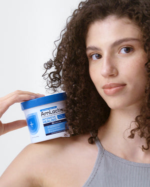 Young woman with brown curly hair holding AmLactin Intensive Healing Cream Tub on her shoulder with one hand. Woman is wearing a grey tank top with a light grey background.
