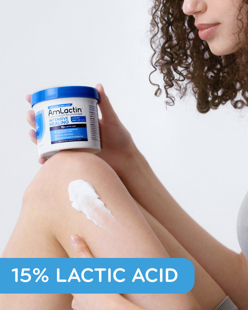 Curly haired woman sitting, holding AmLactin Intensive Healing Cream Tub on her knee. Cream swatch on her knee. 15% Lactic Acid callout flag in blue on bottom of image.