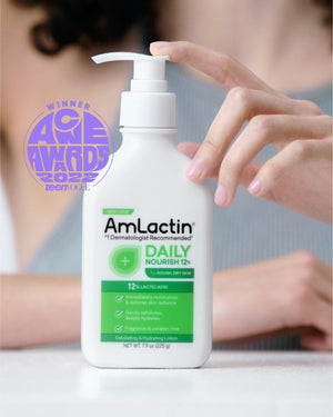 Dark, curly-haired woman wearing a medium grey tank top out of focus holding AmLactin Daily Nourish 12% Lotion 7.9oz Pump Top Bottle on a white surface.Woman's fingers perched on pump top of AmLactin Daily Nourish 12% 7.90 oz bottle. Circular, purple Teen Vogue Acne Awards 2022 badge next to bottle. 