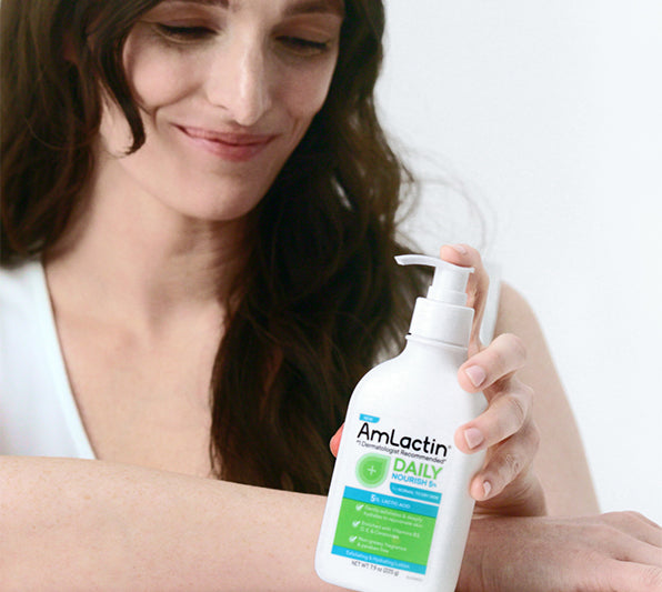 Smiling, dark-haired woman in a white tank top with a light grey background holding AmLactin Daily Nourish 5% Pump Top bottle and resting on a white surface.